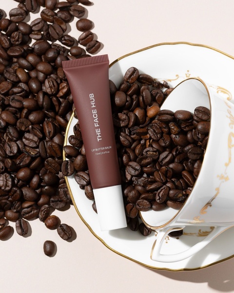 A high-quality, top-down product photography shot of a lip butter balm placed inside a porcelain teacup filled with coffee beans.