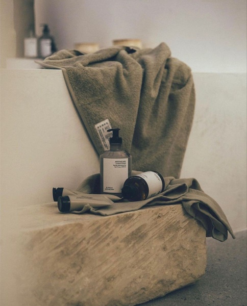 A minimalist, high-end photographic composition featuring apothecary conditioner and toiletries placed on a rustic stone surface with a soft, natural towel backdrop.