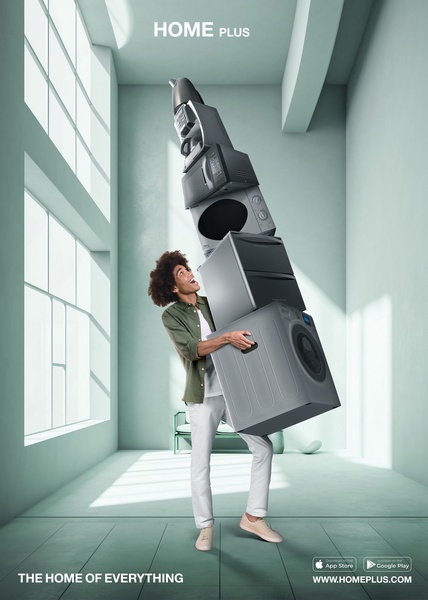 A man joyfully balances a tall, improbable stack of various home appliances to demonstrate the breadth of products available.