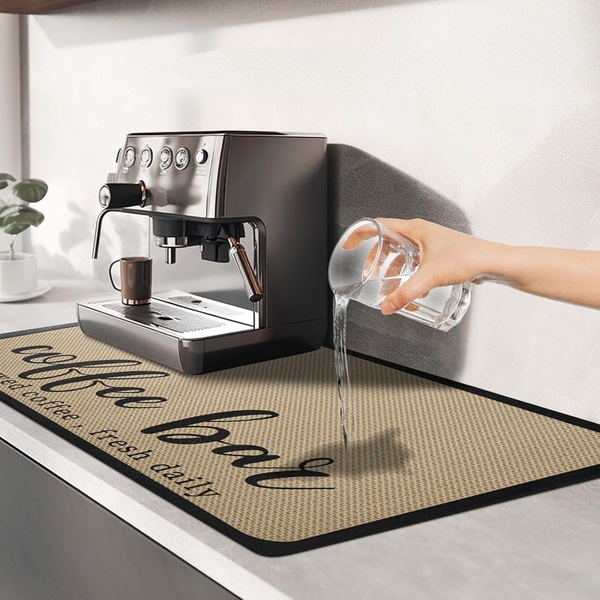 A high-angle studio shot showing water being poured onto a coffee bar mat next to an espresso machine to prove its absorbency.