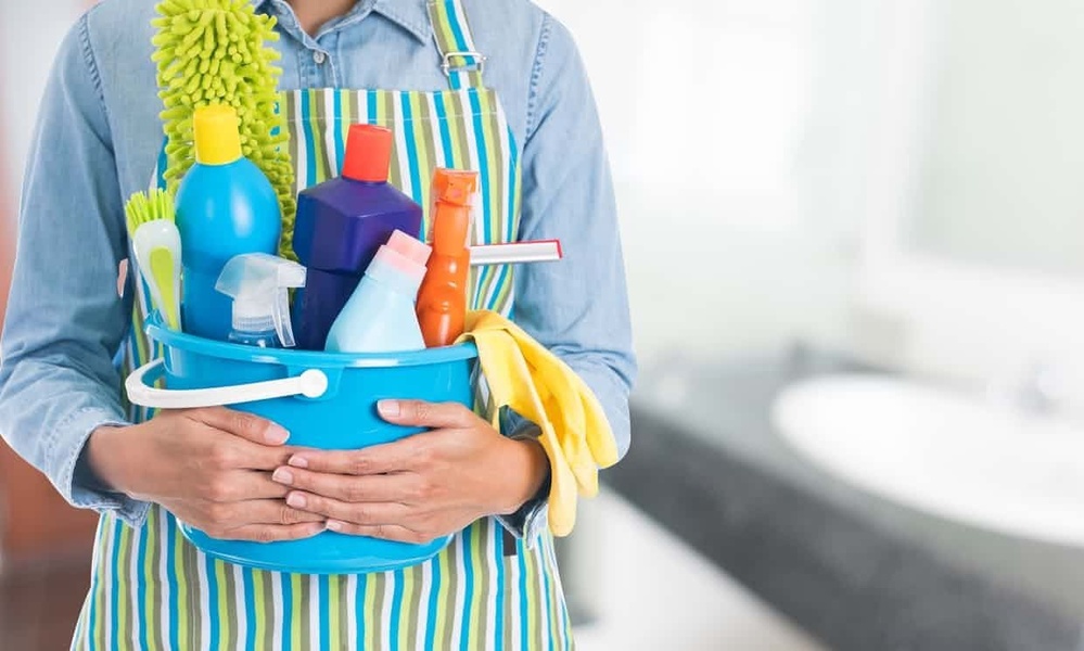 A close-up photograph of a person in an apron holding a blue bucket filled with various colorful cleaning bottles, sponges, and tools, set against a blurred bathroom backdrop.