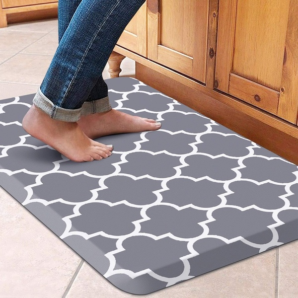 A close-up shot of feet standing on a grey and white geometric anti-fatigue mat placed in front of wooden kitchen cabinets.