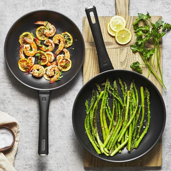 This top-down square shot features two non-stick pans side-by-side, one with sauteed shrimp and lemon, the other with asparagus, emphasizing ease of use and meal prep quality.
