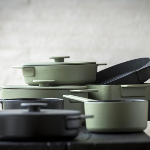 A refined product hero shot featuring a stack of muted olive green and matte black cast iron pots and pans on a dark, rustic surface.