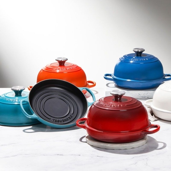 A high-quality 1:1 square product hero shot featuring a collection of enameled cast iron pots in multiple vibrant colors arranged in a clean studio setting.
