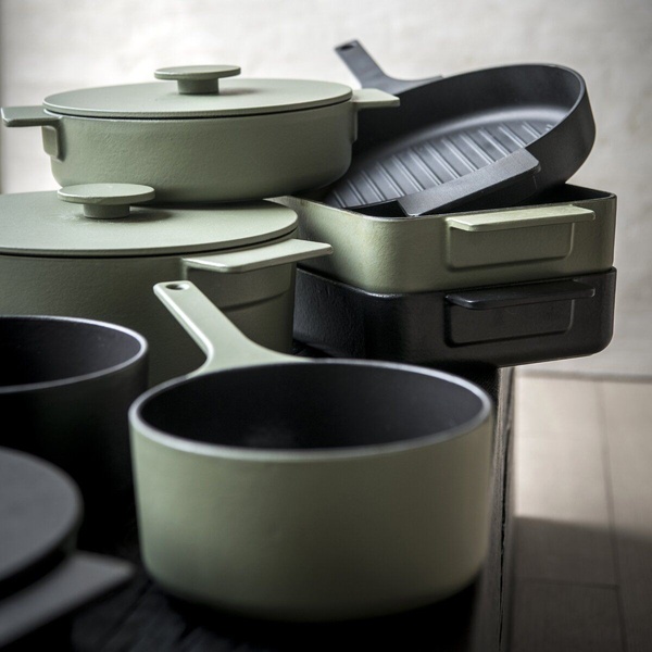 A clean, studio-lit display of stacked matte-finish cast iron pots and pans in sage green and black, emphasizing architectural design and premium quality.