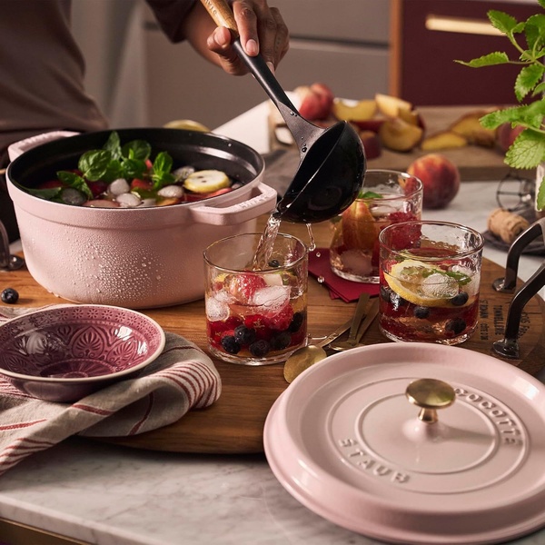 A high-quality lifestyle shot featuring a light pink Dutch oven being used to serve a summer fruit punch at a table.