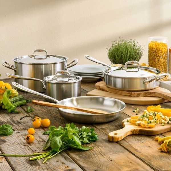 A high-quality lifestyle shot featuring a complete stainless steel cookware set arranged on a rustic wooden table with fresh produce.
