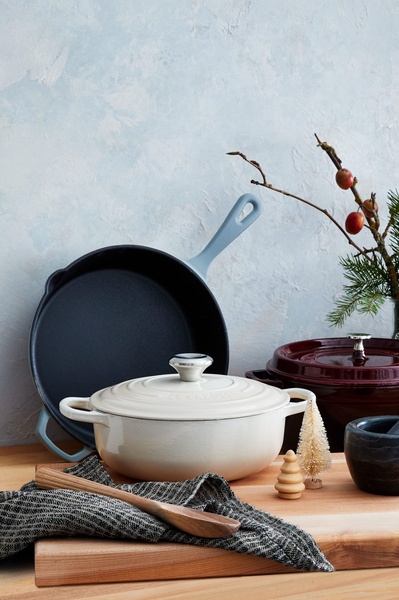 A high-quality studio shot of enameled cast iron cookware featuring a Dutch oven, skillet, and natural decorative elements on a wooden surface.