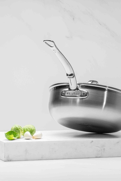 A high-end stainless steel pan is centered against a clean marble background with minimal organic food elements, emphasizing premium quality.