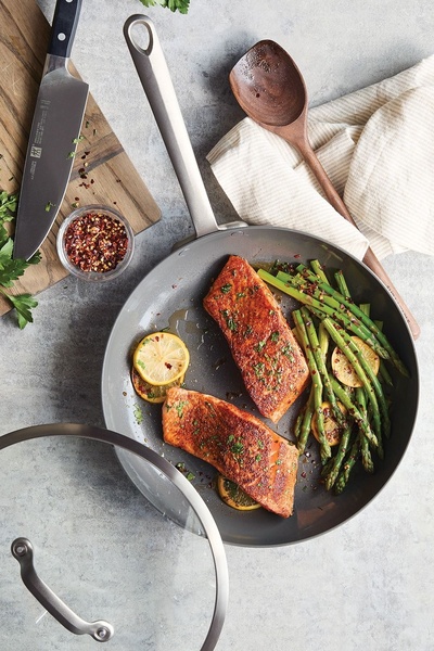 A top-down view of a high-quality nonstick skillet featuring seared salmon, asparagus, and lemon slices, set alongside kitchen tools on a neutral stone background.