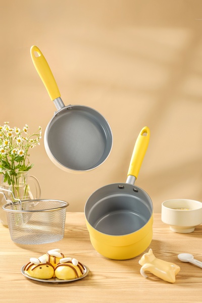 A clean, bright studio shot featuring two yellow, non-stick saucepans floating against a soft beige background with kitchen-themed props.
