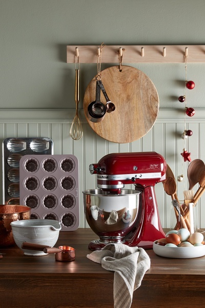 A high-end product hero shot featuring a red stand mixer on a wooden countertop, accented with seasonal baking props in a soft, natural kitchen setting.