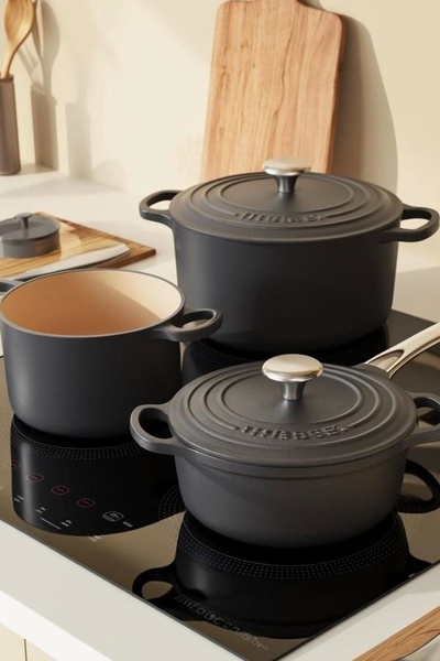 This creative showcases a high-end cast iron cookware set arranged on an induction stovetop, utilizing a neutral, warm color palette to convey luxury.