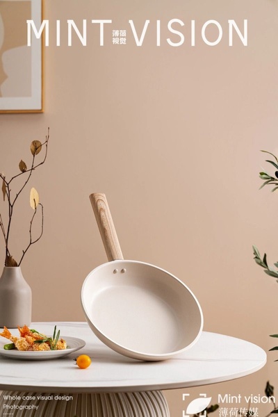 A clean, studio-quality photograph of a beige non-stick frying pan with a wooden handle, styled in a minimalist kitchen setting with a plate of appetizers.