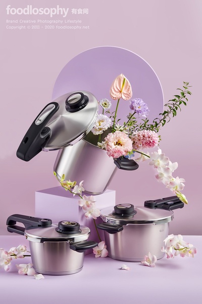A high-end product photography creative featuring stainless steel pressure cookers arranged with delicate pastel flowers against a soft lavender backdrop.
