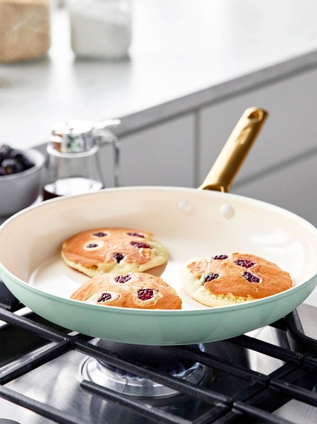 A close-up, high-key photograph of a pastel green frying pan on a gas stove, cooking three fluffy pancakes with blackberries.