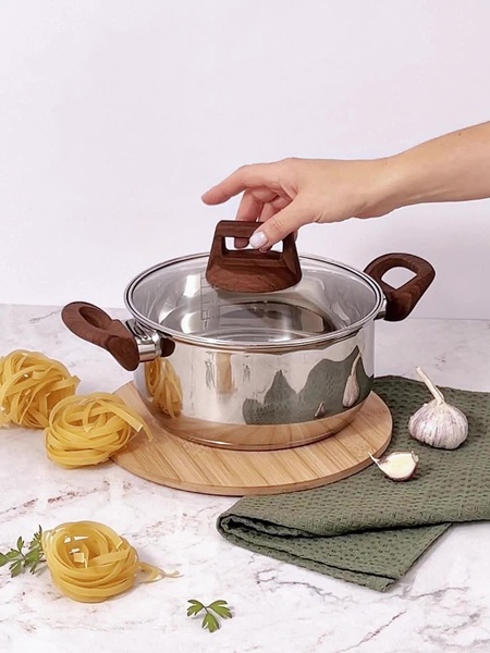 A high-quality lifestyle shot of a polished stainless steel cooking pot placed on a bamboo board with natural ingredients like pasta and garlic.
