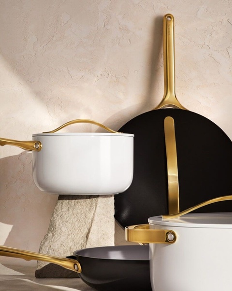 A high-end product photograph featuring white and black cookware pieces with gold handles arranged against a textured stone backdrop.