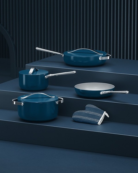 A high-end studio display featuring a premium cookware set, including a saucepan, frying pan, and dutch oven, arranged on tiered platforms in a consistent deep blue finish.