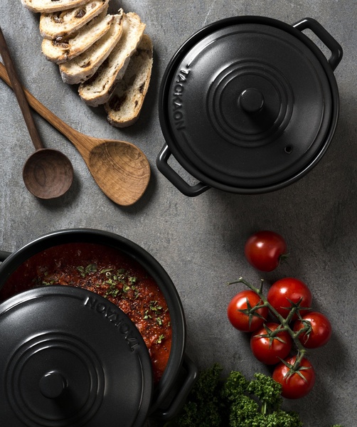 An elegant, high-contrast flat lay photograph showcasing a black cast iron Dutch oven alongside fresh ingredients like tomatoes and crusty bread.