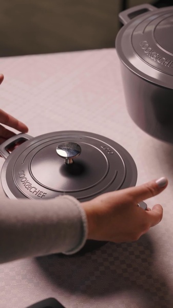 A close-up, high-quality shot focusing on the matte finish and metallic lid handle of a premium grey cast iron dutch oven held by human hands.