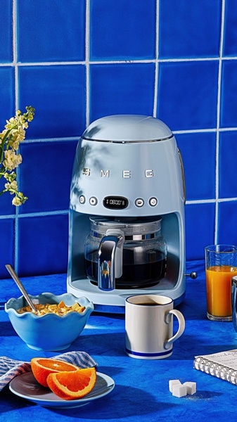 A high-quality 9:16 vertical shot of a light blue Smeg coffee maker positioned on a vibrant blue tiled background with a breakfast setup.