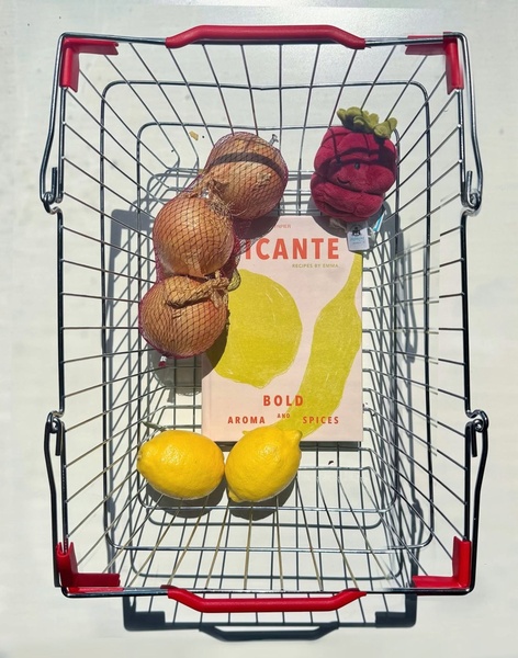 A top-down view of a cookbook inside a shopping basket with fresh ingredients like onions and lemons.