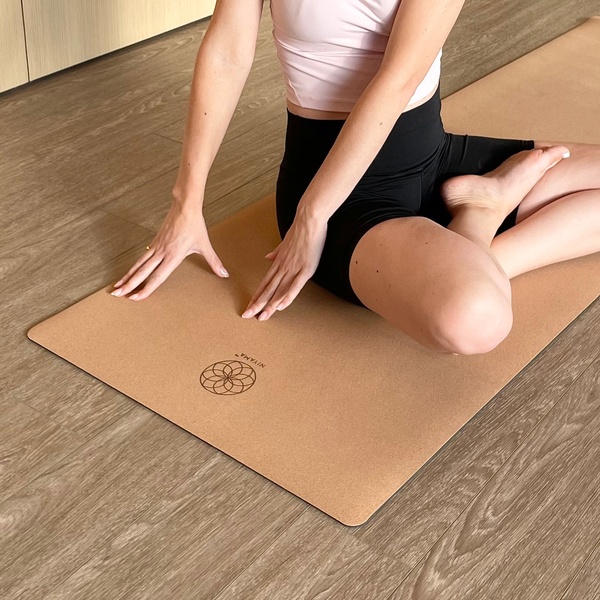 A close-up, top-down shot of a person sitting on a cork yoga mat, focusing on the natural texture and branded logo.