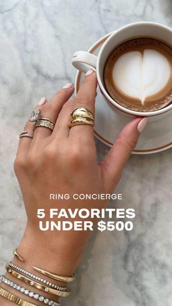 A high-angle, lifestyle close-up showing a hand adorned with multiple gold rings holding a morning latte, highlighting a curated jewelry collection.