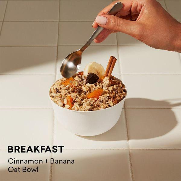 A clean, minimalist 1:1 image featuring a hand holding a silver spoon over a bowl of oatmeal topped with banana slices, cinnamon sticks, and dried apricots.