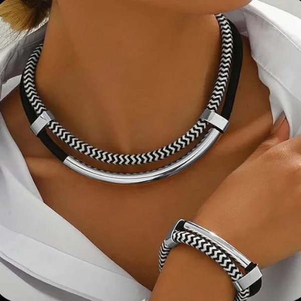 A close-up, high-contrast product shot focusing on a chevron-patterned necklace and matching bracelet on a model wearing a crisp white shirt.