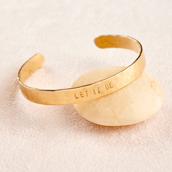 A close-up studio shot of a gold bracelet engraved with 'LET IT BE' resting on a smooth stone, emphasizing a clean and serene aesthetic.