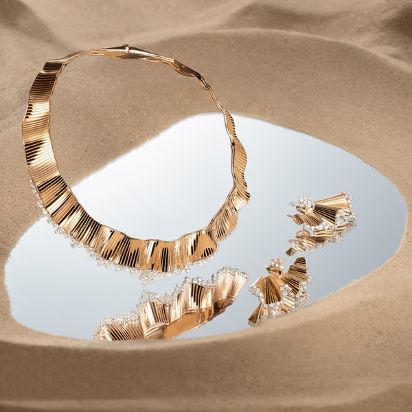 A high-end studio shot of a gold necklace and matching earrings placed on a circular mirror atop soft, textured sand.