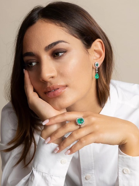 A close-up photograph of a woman wearing emerald and diamond earrings and a matching ring, using a soft, neutral-toned professional studio lighting setup.