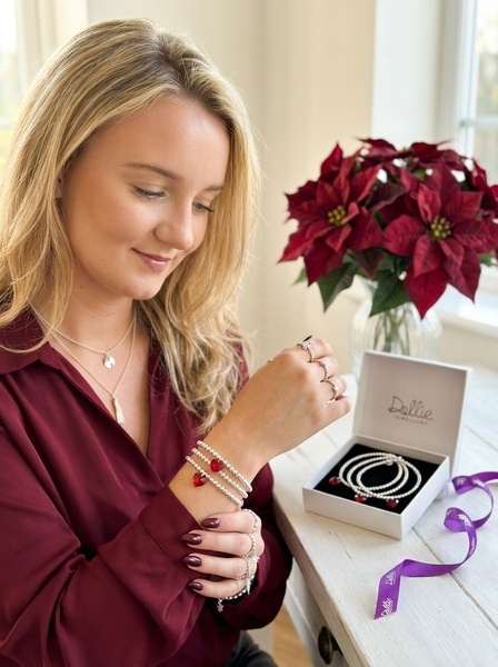 A lifestyle shot showing a woman wearing beaded bracelets with red heart charms, set against a festive backdrop with a holiday poinsettia and gift box.