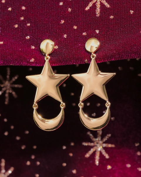 A close-up product shot featuring elegant gold star and crescent moon earrings displayed against a deep red velvet backdrop with glitter accents.
