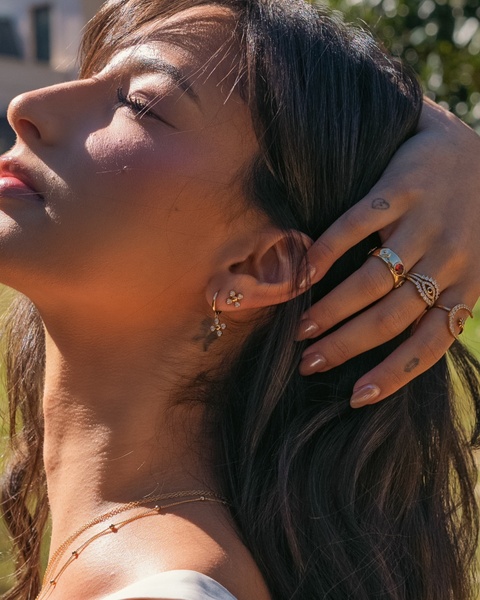 A close-up shot of a model wearing delicate jewelry, featuring focus on earrings and stacked rings with a warm, sun-drenched aesthetic.