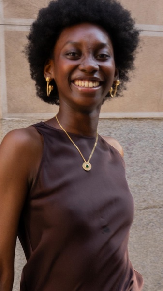 A close-up portrait of a woman wearing a delicate gold chain necklace with a small green pendant and matching gold hoop earrings.