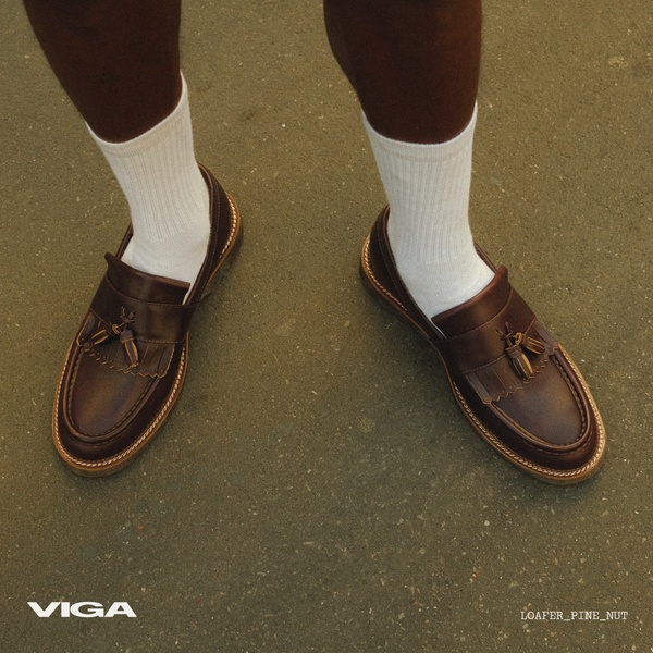 A top-down view of a pair of brown leather tassel loafers worn with white socks, set against a simple asphalt background.
