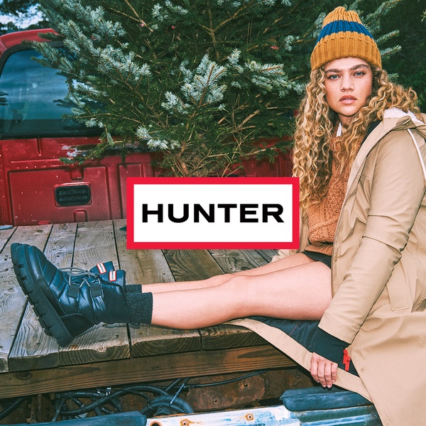 A lifestyle shot featuring a model in winter apparel wearing branded ankle boots, seated on a truck bed near a pine tree to evoke a seasonal feeling.