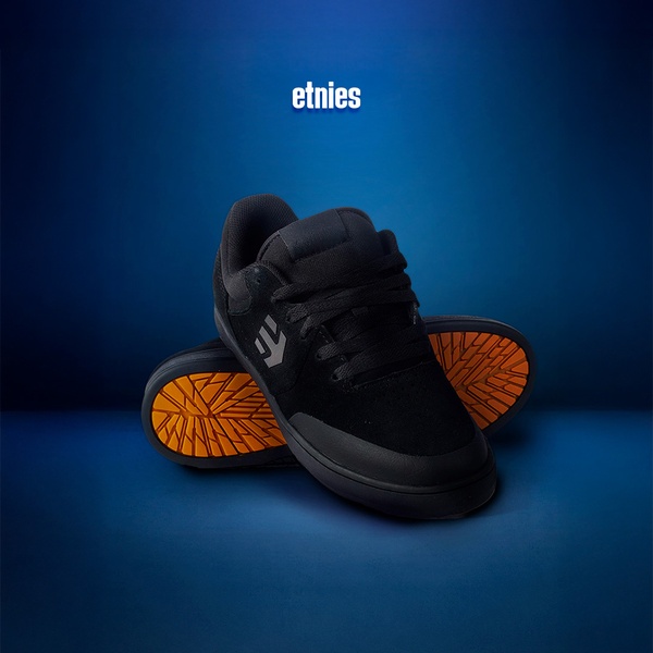 A clean, studio-lit photograph of a black skate shoe resting on its side, emphasizing its orange tread pattern against a deep blue background.