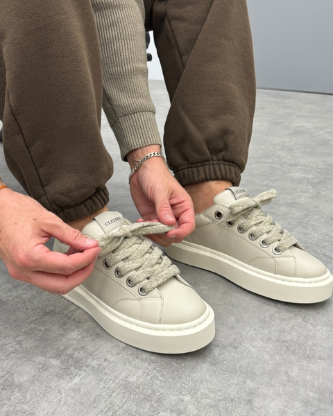 A close-up, first-person perspective showing a pair of clean beige sneakers while a hand adjusts the laces, emphasizing product texture and design quality.