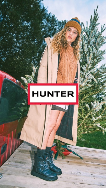 A tall model standing in a flatbed truck wearing seasonal winter attire, including a long beige coat and dark winter boots.