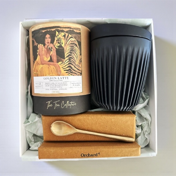 A high-end, top-down flatlay shot of a tea brand gift set featuring Golden Latte mix, a reusable travel cup, and a brass spoon.