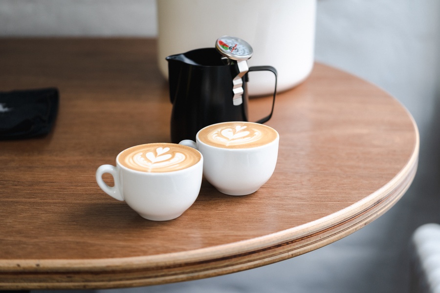 A high-quality, top-down-angled close-up of two cups of latte art on a wooden surface, featuring a coffee thermometer in a black pitcher.