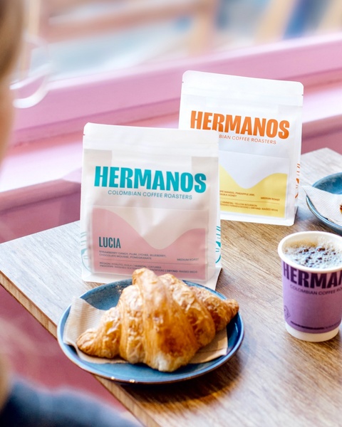 A clean, high-contrast product shot featuring coffee bean bags alongside a morning pastry and a cup of freshly brewed coffee.