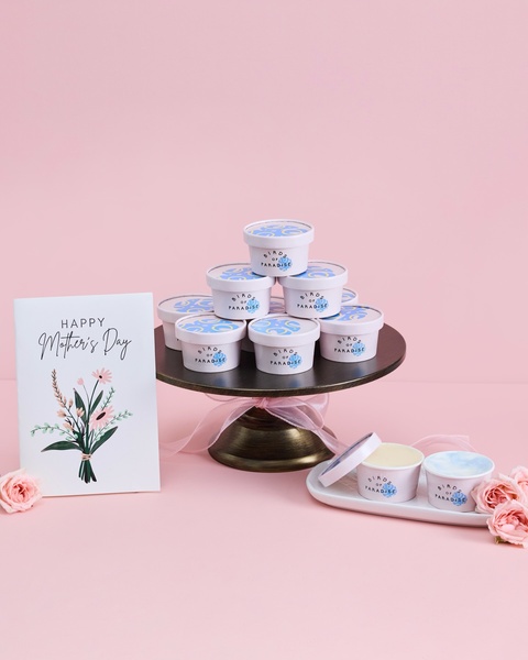 A professionally styled product hero shot featuring stacked ice cream tubs on a cake stand next to a Mother's Day greeting card and fresh roses.