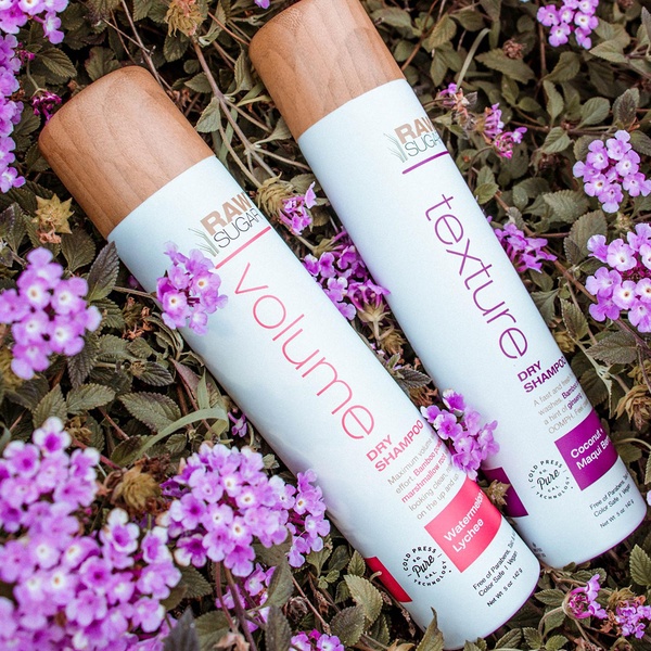 A top-down 1:1 flat lay of two Dry Shampoo bottles placed amidst purple lantana flowers to emphasize natural ingredients and product volume.
