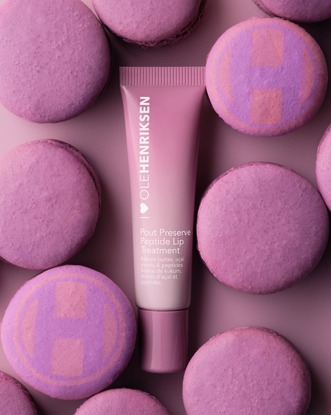 A close-up studio shot featuring a Pout Preserve Peptide Lip Treatment tube surrounded by matching purple macarons.